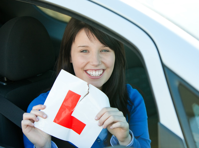 An image depicting a young happy recently passed female student displaying a red Learner plate out of the window of the drivers seat