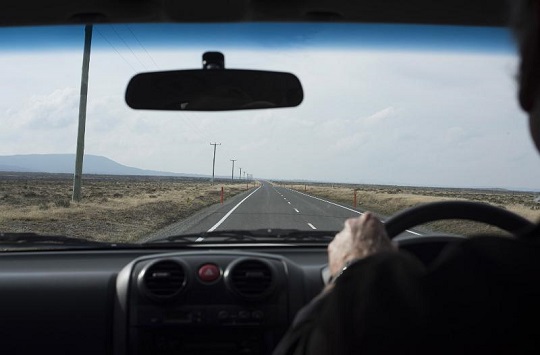An image depicting a Long journey ahead concept with man driving a car in an over the shoulder perspective looking at the long road stretching into the distance through the windscreen