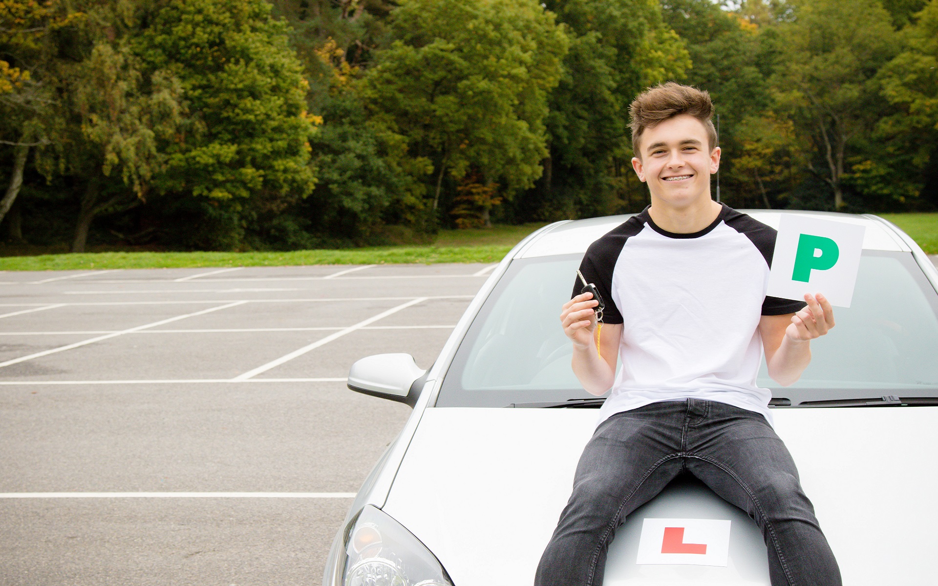 An image depicting a young male holding a 'Passed' sticker plate in his left hand and car keys in his right what sat on vehicle with a red Learner stick label on the bonnet.